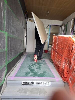 交野市の建築現場で養生シートを確認しながら行う板状資材の荷揚げ作業